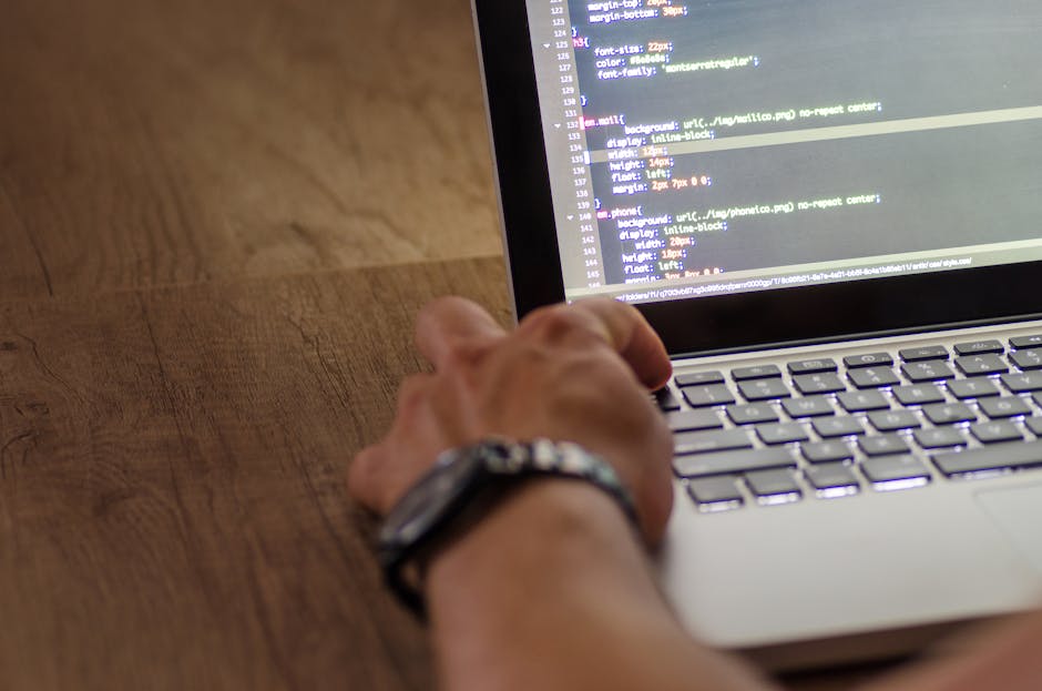 A close-up view of a person's hand wearing a silver wristwatch, working on a laptop placed on a wooden desk. The laptop screen displays lines of HTML or code in a text editor, with the background of t